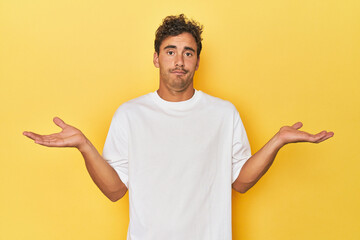 Young Latino man posing on yellow background doubting and shrugging shoulders in questioning...