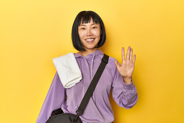 Chinese lady in gym attire, yellow studio smiling cheerful showing number five with fingers.