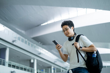 Young Asian millenael man standing in train station or airport while looking smartphone with black...