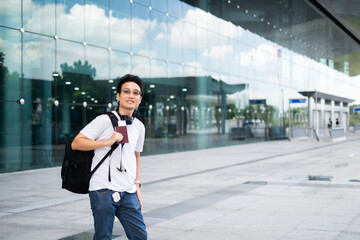 Obraz premium Young Asian millenael man standing with smile in front of air port ready for trip travel alone with passport, black backpack and headphone.