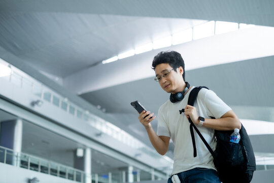 Young Asian millenael man standing in train station or airport while looking smartphone with black backpack and headphone with happiness.
