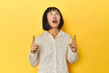 Young Chinese lady, yellow studio background pointing upside with opened mouth.