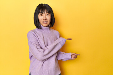 Young Chinese lady, yellow studio background shocked pointing with index fingers to a copy space.