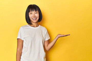 Young Chinese lady, yellow studio background showing a copy space on a palm and holding another...