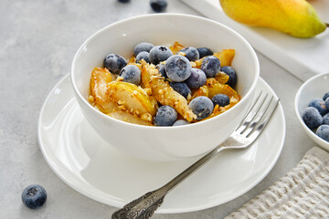 Cottage cheese with pieces of pears, nuts and blueberries in a white cup on a light-gray background. Delicious and healthy breakfast