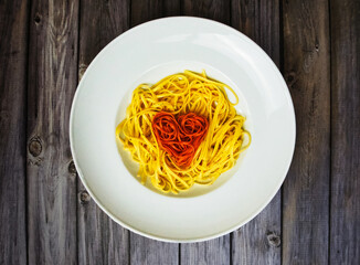 Rote Linguine herzförmig auf gelben Linguine auf einem weißen Teller angerichtet, horizontal, hölzerner Hintergrund 
