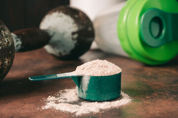 Chocolate whey protein powder in measuring spoon, old rusty dumbbell and shaker on dark background....