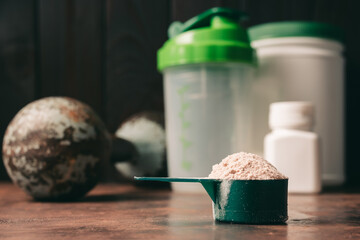 Chocolate whey protein powder in measuring spoon, old rusty dumbbell, shaker, plastic bottle on...