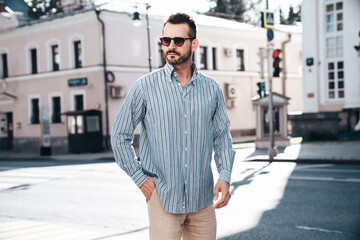 Portrait of handsome confident stylish hipster lambersexual model. Sexy modern man dressed in blue shirt and trousers. Fashion male posing on street background in Europe city at sunset. In sunglasses