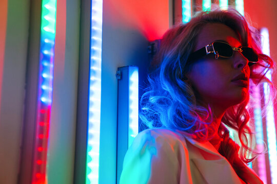 Young beautiful hipster woman in street style fashion concept. Hot model wearing white suit and sunglasses. Female posing in red and blue neon light in studio interior