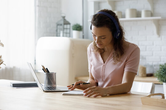 Focused millennial student woman in wireless headphones writing learning notes, reading summary at laptop computer in home kitchen, listening to audio lecture, webinar, attending virtual class
