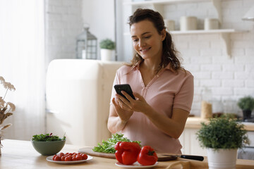 Happy millennial food blogger girl taking picture of fresh vegetables for salad on kitchen table, shooting healthy ingredients on smartphone, reading online recipe, texting message on internet