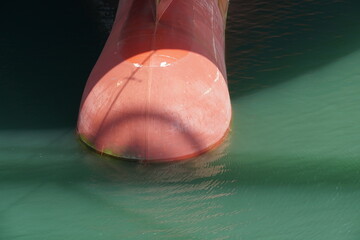 Red bulbous bow of container vessel in detail view, which is half submerged in green colored water....