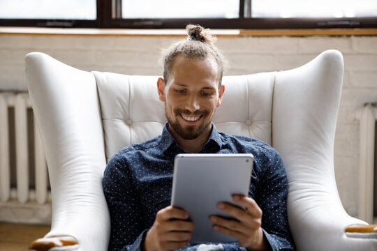 Happy millennial guy using digital device at home, making video call on tablet, reading ebook, browsing internet, social media, shopping with online app, resting in comfortable armchair at home