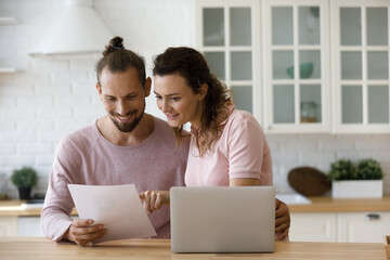 Happy satisfied millennial married couple reading paper document at laptop in home kitchen,...