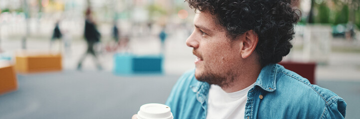 Close-up portrait of young bearded man in denim shirt sitting in wireless headphones and drinking coffee on busy street modern city background. Profile of man drinking coffee