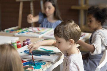 Obraz premium Adorable multiracial children sit at table drawing with paints on canvas. Creative hobby, favourite activity, painting class to nurture kids creativity and imagination, to improve fine motor skills
