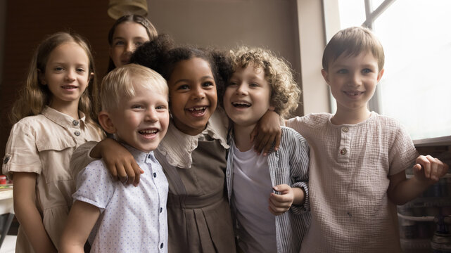 Cute six happy kids pose in art-studio, hugging enjoy creative hobby and friendship after painting classes. Portrait of cheerful multiethnic alpha generation girls and boys laughing looking at camera
