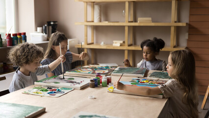 Group of cute little serious boy and multiethnic girls sit at table in art-studio engaged in artistic class, painting pictures with paints on canvas. Children development, creative hobby, day-care