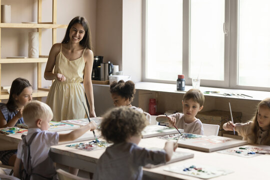 In cozy creative studio group of multi-ethnic children attend art class led by young artist woman teach cute preschoolers to paint pictures on canvas. Kids development, educational courses, hobbies