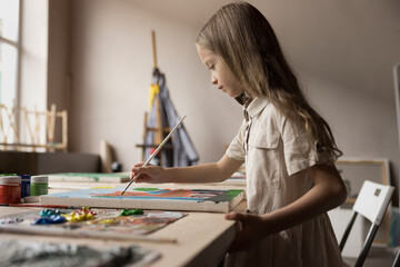 Serious focused little kid girl holding paintbrush, makes strokes on canvas, painting with acrylic...