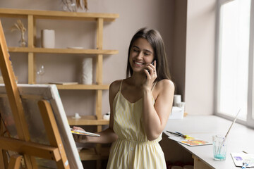 Happy young painter woman standing in art studio near easel holds palette with paints and talking, lead conversation by work or personal use modern smart phone. Mobile connection, hobby, communication