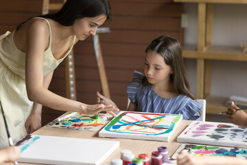 Young artist woman teach little girl during class in art-studio, children sit at table enjoy painting pictures of canvas with paints, lesson led by caring attractive female painter in workshop. Hobby