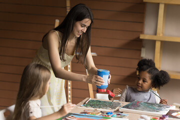 Young female painter, art-class teacher teaching talented creative children, helps with painting, add paints, enjoy communication with preschoolers multiethnic cute kids. Daycare, hobbies, development