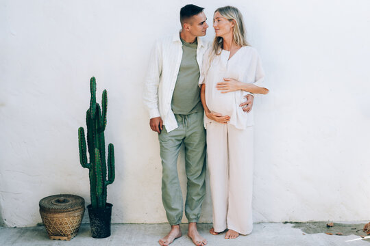 A Married Pregnant Couple Stands Against A White Wall. Tender Love Relationship Of A Man And A Woman