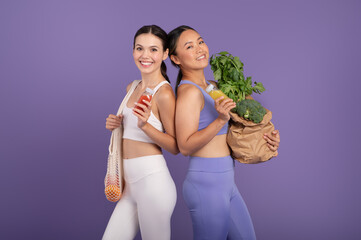 Caucasian and asian women hold smoothies and vegetables, posing over purple studio background