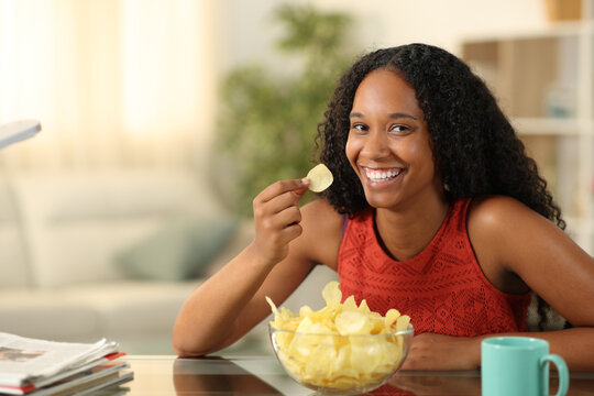 Happy black woman eating potato chips looking at you