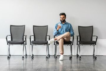Businessman sitting in recruitment waiting room for interview with HR, using cellphone, full length