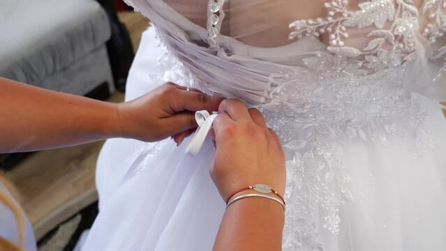 A friend of the bride helps her put on her wedding dress by tying the laces on the back. Preparation for the wedding ceremony.