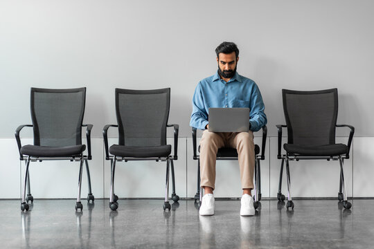 Indian man sitting on chair, waiting for interview and using laptop computer, full length, free copy space