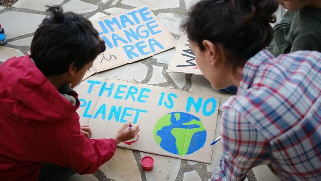 A mother and her two children prepare colorful protest signs against climate change and pollution using paintbrushes