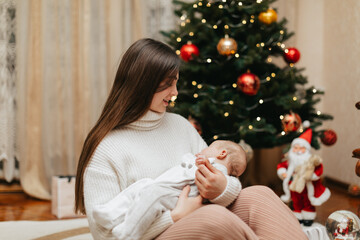 Infant baby girl in a Christmas romper is sleeping in her mother’s hands at home on Christmas Day. Mom is holding a sleeping newborn baby in her arms near the Christmas tree at home on Christmas. 