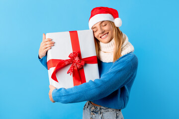 Excited caucasian woman in Santa hat holding and hugging big wrapped gift box over blue studio background
