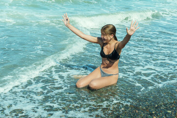 Happy emotional teenage girl sits in shallow water in foamy stormy sea waves on a sunny warm summer day.