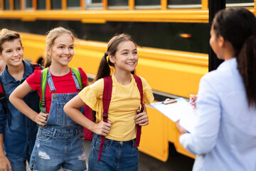 Happy children boarding school bus while black female teacher updating check list