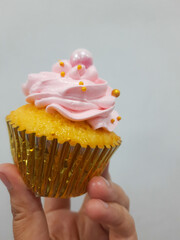 Yellow cupcake with pink cream, edible pearls and gold cupcake liner. Hold by hand, isolated on white background