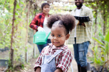 Happy cute litter African Asian mixed-race girl with curly hair working in farm together with parents, child with mother and father at behind, happy family in horticulture garden at countryside.