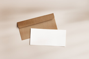 Blank paper card mock up and craft envelope on beige background. White empty letter or invitation on table, sunlight, beautiful shadow. Minimal modern lifestyle. Flat lay, top view, copy space.