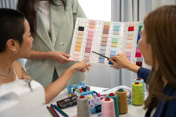 Female designer learning to make dress at designer school. They pointing to pantone together. Seamstress teaching asian woman to sew in class. - obrazy, fototapety, plakaty