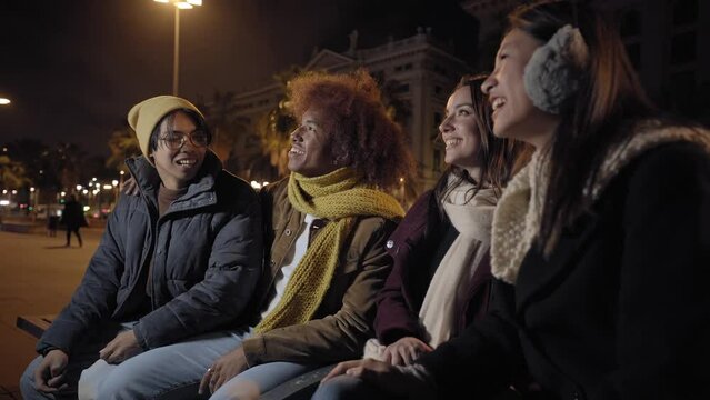 Generation z friends chatting and laughing sitting in city square at night. Group of excited people hanging out together. Multiracial couples meeting having fun. Positive young adult relationships.