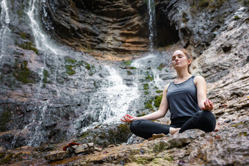 Young woman practicing yoga, sitting in om pose on the rock near the beautiful waterfall. Relax, nature, and wellbeing concepts.