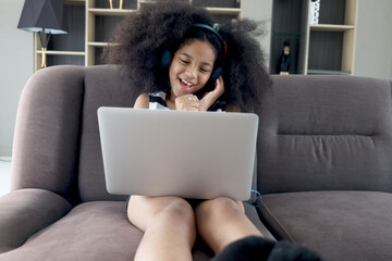 Naklejka premium Happy smiling beautiful teenage girl with curly hair wearing headphones, using laptop computer for practicing to sing song while sitting on sofa in home living room.