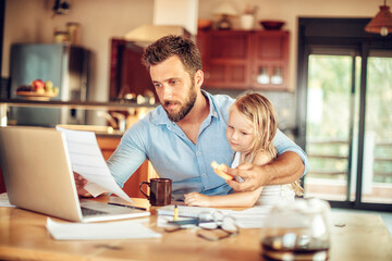 Concerned young father going over bills while holding his daughter in the kitchen - obrazy, fototapety, plakaty