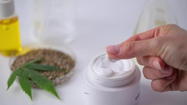 Female hand checking cannabis hemp cream. Container with hemp skin care cream, CBD oil, THC tincture and cannabis leaves. Cannabis medical herbal extract. Organic skin care CBD oil cream.