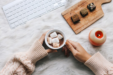 Female hand holding a cup of chocolate marshmallow near computer keyboard, brownies cake on wooden...
