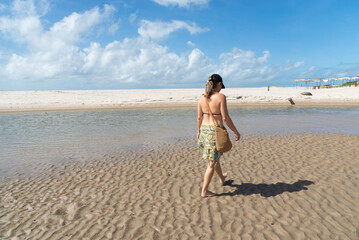 Fototapeta na wymiar A beautiful woman in a bikini walking on the wavy sand of a beach. Blue sky and clouds in the background. Taquari Beach in Valenca, Brazil.